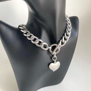 K&ouml;nnte beinhalten: Eine silberne Kettenkette mit einem gro&szlig;en, klobigen Gliederdesign. Die Kette hat einen Toggle-Verschluss und einen herzf&ouml;rmigen Anh&auml;nger.