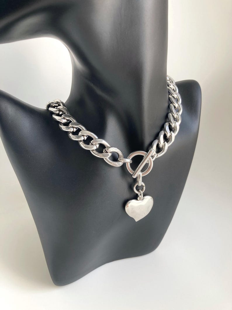 K&ouml;nnte beinhalten: Eine silberne Kettenkette mit einem gro&szlig;en, klobigen Gliederdesign. Die Kette hat einen Toggle-Verschluss und einen herzf&ouml;rmigen Anh&auml;nger.