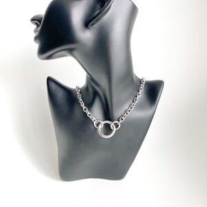 May include: A silver chain necklace with a large, round, silver pendant. The necklace is on a black mannequin.