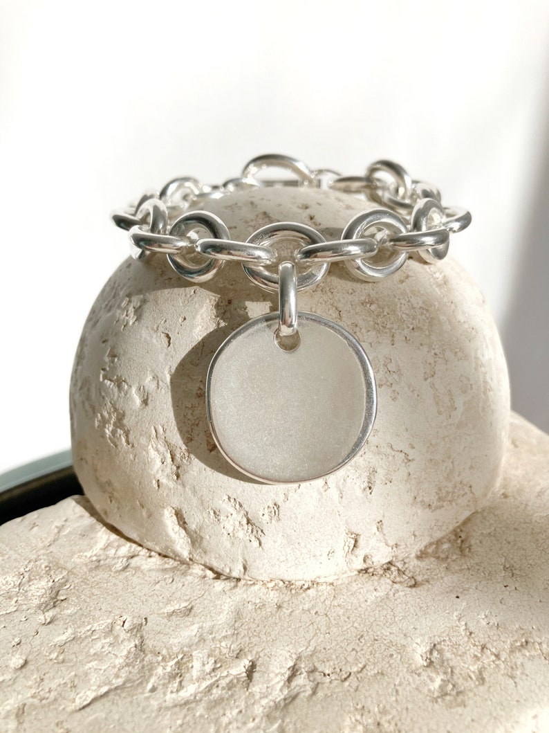 Peut inclure: Un bracelet en argent avec une grosse breloque ronde en argent. Le bracelet est pos&eacute; sur une surface blanche textur&eacute;e.