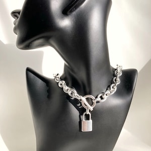 Puede incluir: Un collar gargantilla de cadena plateada con un colgante de candado. El collar está en un maniquí negro.