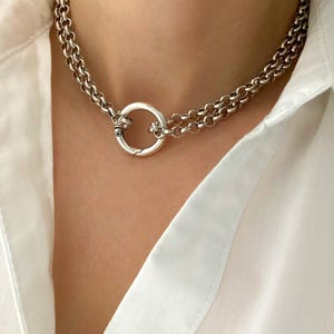Puede incluir: Un collar plateado con un diseño de doble cadena. El collar tiene un cierre circular grande en el centro. Las cadenas están hechas de eslabones redondeados. El collar se usa sobre una camisa blanca.