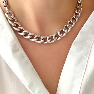 Peut inclure: Un collier chaîne argenté avec de gros maillons. Le collier est porté sur une chemise blanche. La chaîne est un bijou audacieux, parfait pour ajouter une touche d'originalité à n'importe quelle tenue.