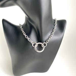 May include: A silver chain necklace with a large, round, silver pendant. The necklace is on a black mannequin.
