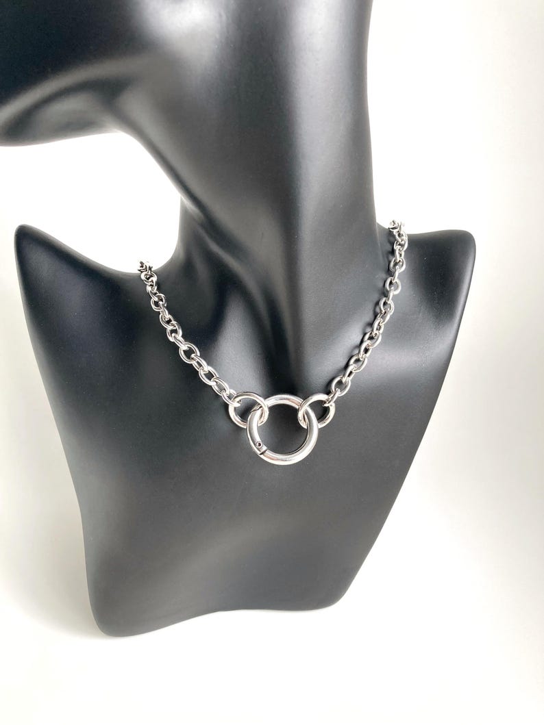May include: A silver chain necklace with a large, round, silver pendant. The necklace is on a black mannequin.