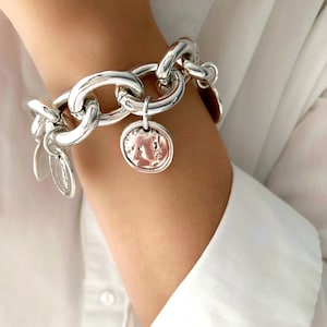 Könnte beinhalten: Ein silbernes Kettenarmband mit großen ovalen Gliedern. Das Armband hat einen Münzanhänger mit Gesichtsdesign und mehrere kleinere Anhänger. Das Armband wird am Handgelenk getragen, mit einem weißen Hemd im Hintergrund.