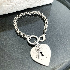 May include: A silver chain bracelet with a heart-shaped charm featuring a keyhole and a key. The charm is attached to the chain with a silver ring.