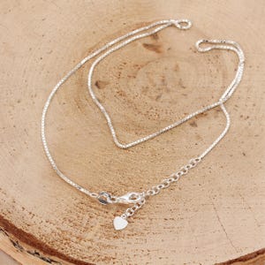 May include: A delicate silver necklace with a box chain design. The necklace features a heart-shaped charm and an adjustable clasp. The chain is displayed on a light brown wooden surface.