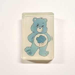 May include: A blue Care Bear with a cloud and raindrops on its belly. The bear is standing with its arms crossed and has a frown on its face. The bear is in a clear plastic cube.