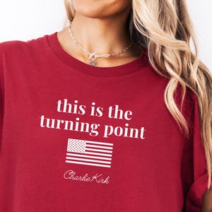 May include: A maroon t-shirt with the text "this is the turning point" above a graphic of the American flag and the name "Charlie Kirk" in white. The shirt has rolled-up sleeves.