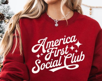 Può includere: Felpa rossa con testo bianco che recita "America First Social Club" in un font retrò con stelle scintillanti.