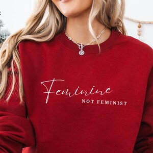 May include: A burgundy sweatshirt with the words "Feminine" in cursive script above "NOT FEMINIST" in a sans-serif font. The sweatshirt is a solid color and has a crew neck.