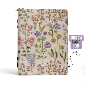 Op de afbeelding: Een witte leren notitieboekomslag met een geborduurd bloemenmotief. De borduurwerk toont verschillende bloemen in tinten roze, paars, geel en groen. De tekst "CUSTOM NAME HERE" is zichtbaar in een paarse doos aan de rechterkant van de omslag.