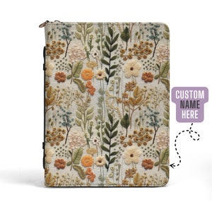 Könnte beinhalten: Ein Notizbuch mit floralem Stickdesign und Reißverschluss. Der Einband hat einen cremefarbenen Hintergrund mit einem sich wiederholenden Muster aus gestickten Blumen und Blättern in Grün-, Orange- und Cremetönen. Der Text "CUSTOM NAME HERE" steht rechts.
