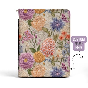 Puede incluir: Una cubierta de cuaderno de cuero blanco con un diseño de bordado floral. Las flores son en tonos de rosa, morado, amarillo y azul. La cubierta tiene una cremallera y una correa negra. El texto "CUSTOM NAME HERE" es visible en la esquina superior derecha de la imagen.