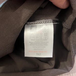 May include: Close-up of a brown garment tag with care instructions and the text "100% COTTON." The tag also includes the text "KEEP AWAY FROM FIRE" and the code "ML285/987."