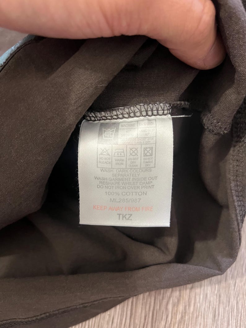 May include: Close-up of a brown garment tag with care instructions and the text "100% COTTON." The tag also includes the text "KEEP AWAY FROM FIRE" and the code "ML285/987."