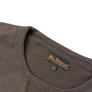 May include: Close-up of a brown Ben Sherman crew neck shirt. The shirt has a ribbed neckline and a stitched design on the front. The Ben Sherman label is visible, with the size XL.