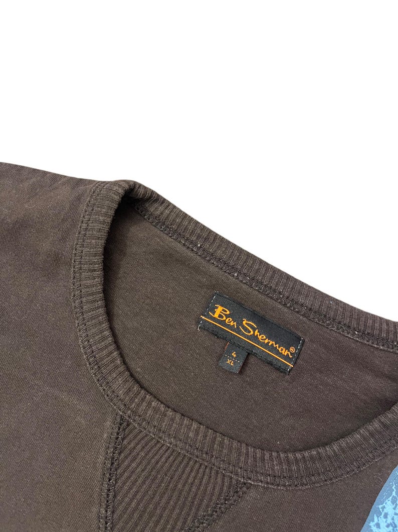 May include: Close-up of a brown Ben Sherman crew neck shirt. The shirt has a ribbed neckline and a stitched design on the front. The Ben Sherman label is visible, with the size XL.