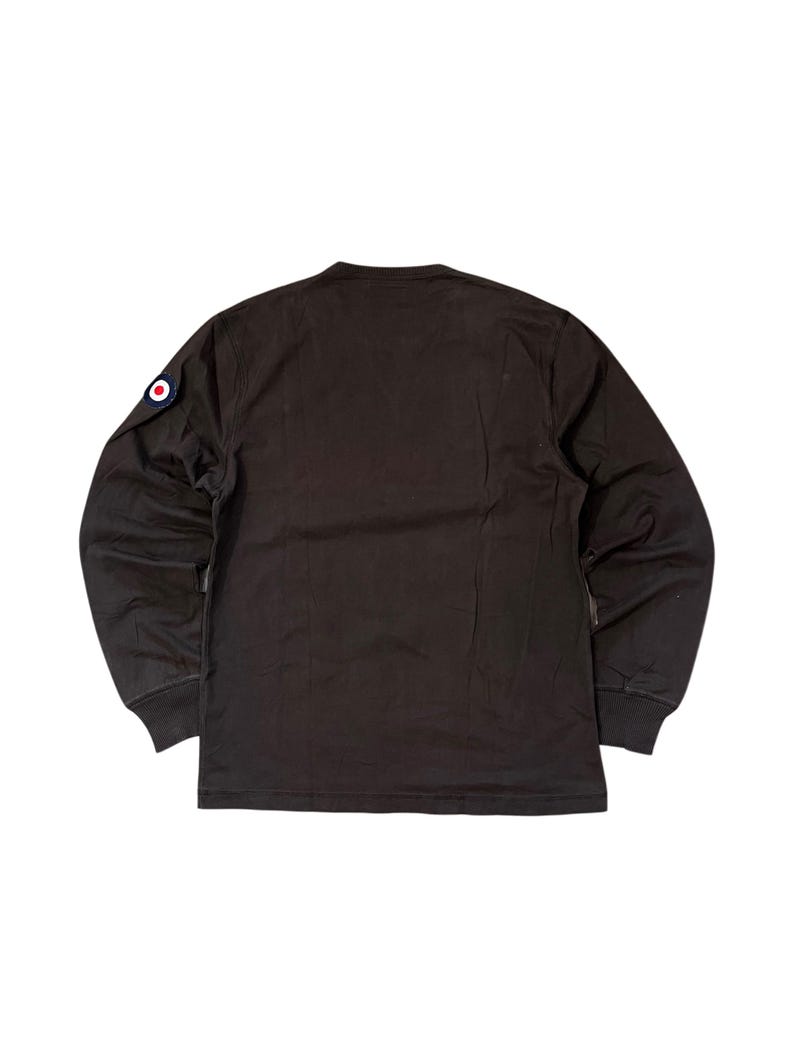 May include: A dark brown long-sleeve shirt with a crew neck. A circular patch with red, white, and blue accents is on the left sleeve. The shirt has ribbed cuffs and a straight hem.