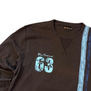 May include: A brown Ben Sherman sweatshirt with the number "63" in light blue, along with the brand name. The sweatshirt has a blue and gray stripe design on the right side.
