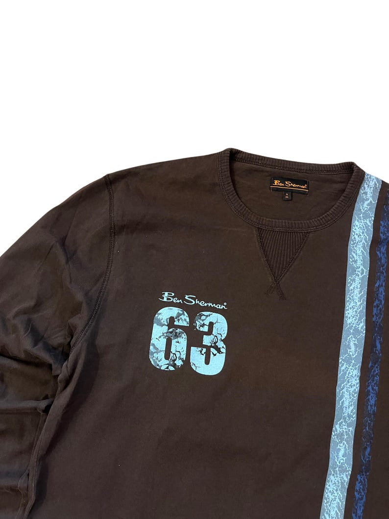 May include: A brown Ben Sherman sweatshirt with the number "63" in light blue, along with the brand name. The sweatshirt has a blue and gray stripe design on the right side.