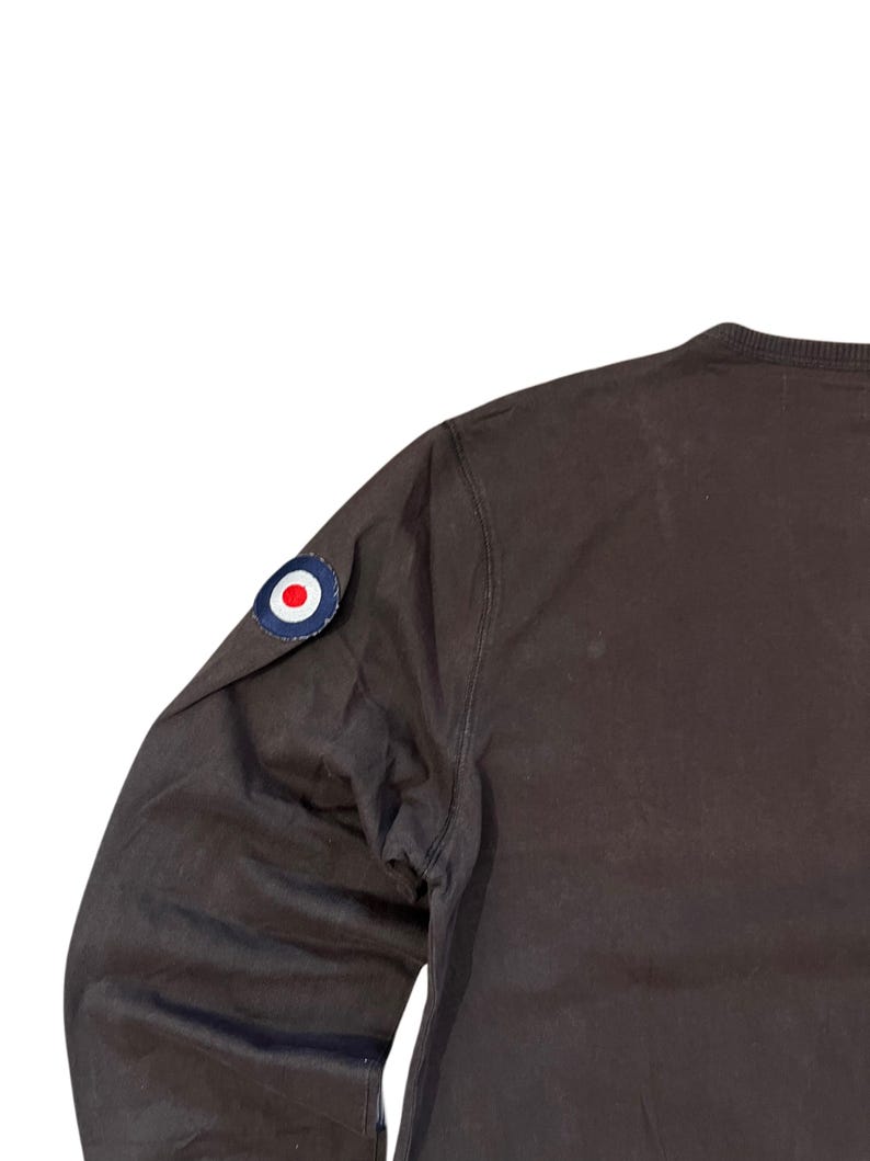 May include: A dark brown long-sleeve shirt with a round patch on the upper left sleeve. The patch is a blue circle with a white border and a red and white center. The shirt is made of a smooth fabric.