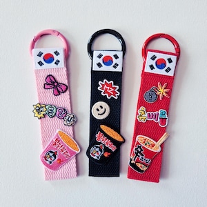 May include: Three keychains with Korean flag accents. One pink keychain features a bow and a ramen patch. A black keychain has a smiley face and a ramen patch. A red keychain has a bomb and ramen patch.