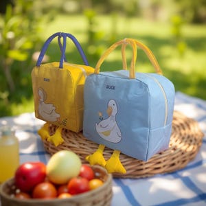 May include: Two insulated lunch bags, one yellow and one blue, each with a duck design and the word "DUCK". The bags have yellow duck feet and handles. A basket of fruit and a drink are in the foreground.