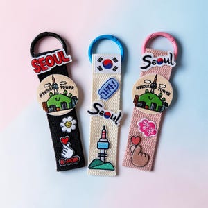 May include: Three fabric keychains with embroidered patches. One is black with a white flower and a hand gesture with a heart. The second is off-white with a Korean flag. The third is pink with a hand gesture with a heart.