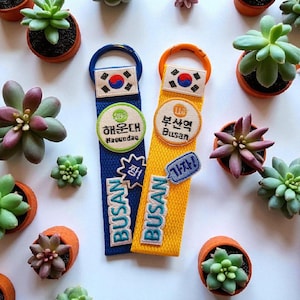 May include: Blue and yellow Busan keychains with Korean flag patches and city names. The keychains are surrounded by small potted succulents. The blue keychain says "Haeundae" and the yellow keychain says "Busan".