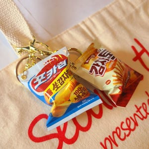 Pode incluir: Dois chaveiros em miniatura de sacos de snacks. Um é azul, branco e vermelho com texto coreano e um gráfico de batata. O outro é bege e vermelho com texto coreano.