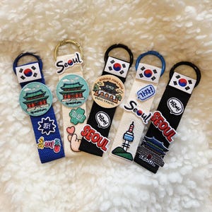 Pode incluir: Cinco chaveiros com o tema Seul. Cada chaveiro apresenta uma fita de tecido com um anel de metal, um patch com texto e imagens coreanas e uma pequena bandeira coreana. Os chaveiros são em cores diferentes, incluindo azul, preto e branco.