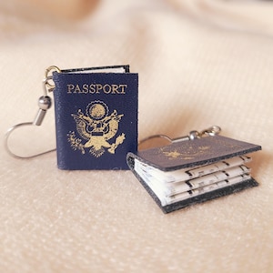 May include: Miniature navy blue passport earrings with silver-tone ear wires. The passport covers feature gold embossed text and a gold emblem. One earring is open, revealing the pages inside. The earrings are on a soft, neutral-toned surface.