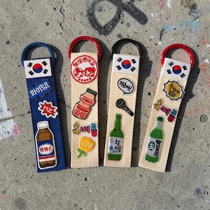 May include: Four fabric keychains with Korean flag designs and various embroidered patches. The patches include images of bottles, a microphone, and text in Korean. Each keychain has a different colored ring: blue, red, black, and red.