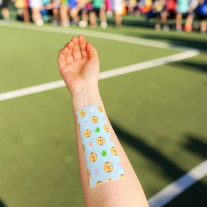 May include: A light blue athletic tape with a repeating pattern of orange tiger faces, green leaves, and pink stars is applied to a person's forearm. The background is a green sports field with white lines.