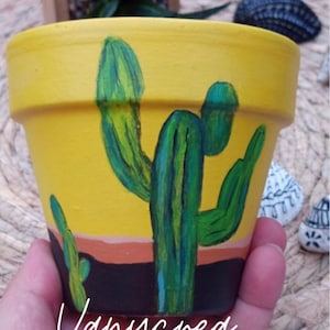 Peut inclure: Un pot de fleurs en terre cuite jaune orné d'un motif de cactus peint à la main en vert. La base du pot présente une bande horizontale orange au-dessus d'une bande noire. Le texte cursif blanc Vanycrea est inscrit sur le bas du pot.