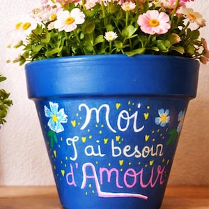 Peut inclure: Un pot de fleurs bleu vif rempli de marguerites roses et blanches. Le pot est peint à la main avec la phrase française "Moi j'ai besoin d'Amour" en lettres blanches et roses, et des motifs floraux.