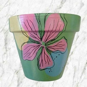 Könnte beinhalten: Ein grüner Terrakotta-Blumentopf mit handbemaltem pink- und blauem Blumendesign.