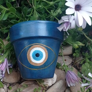 Peut inclure: Un pot de fleurs en terre cuite peint en bleu foncé avec un symbole de mauvais œil peint à la main sur le devant. Le motif se compose d'une pupille noire centrale, d'un iris bleu clair, d'un anneau blanc et d'un contour doré.