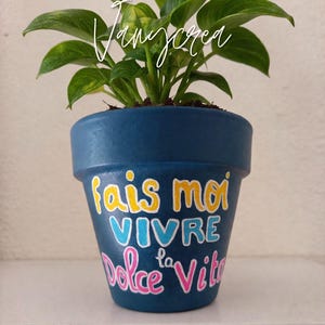 Peut inclure: Un petit pot de plante avec des feuilles vertes dans un pot en céramique peint en bleu foncé. Le pot porte les mots "fais moi vivre la Dolce Vita" en jaune, bleu et rose. Le pot est posé sur une surface blanche.