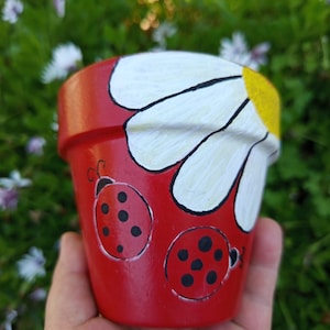 Peut inclure: Pot de fleur rouge peint avec un motif de marguerite blanche et deux illustrations de coccinelles. Le pot de fleur est tenu dans une main sur un fond vert flou.