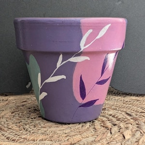 Può includere: Un vaso di terracotta viola e rosa con un disegno dipinto a mano di foglie bianche e viola.