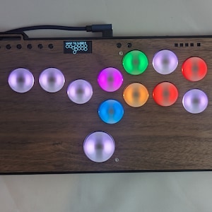 Może przedstawiać: Drewniany kontroler muzyczny z 11 okrągłymi przyciskami, każdy z innym kolorowym światłem. Przyciski są ułożone w półkole. Kontroler ma mały ekran, na którym wyświetlany jest tekst "SUPER 16 PLAYER".