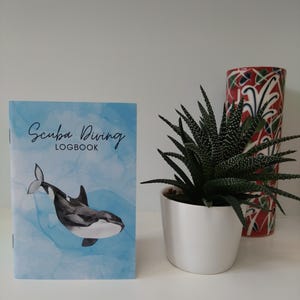 Puede incluir: Un libro de registro de buceo azul y blanco con el texto "Scuba Diving Logbook" y una ilustración en blanco y negro de una orca nadando en el agua.