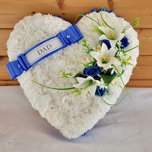 May include: White heart-shaped floral arrangement with blue ribbon and white lilies. The ribbon has the word "DAD" printed on it.