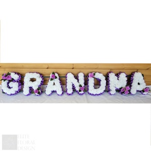 May include: White floral letters spelling out the word "GRANDMA" with purple flowers and ribbon accents.