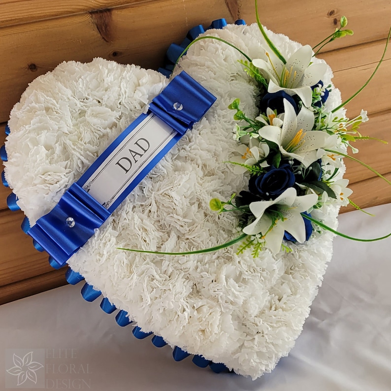 May include: White heart-shaped floral arrangement with blue ribbon and a white card with the word "DAD" on it. The arrangement is decorated with white lilies and blue roses.