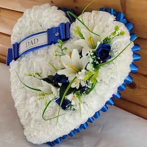 May include: White heart-shaped funeral wreath with blue ribbon and white lilies, blue roses, and greenery. The ribbon has the word "DAD" written on it.