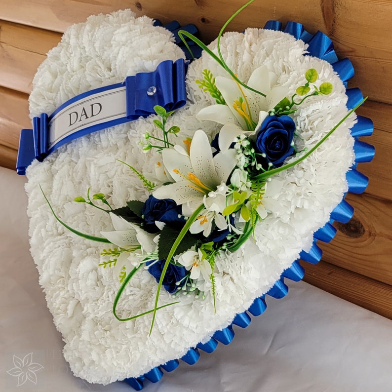 May include: White heart-shaped funeral wreath with blue ribbon and white lilies, blue roses, and greenery. The ribbon has the word "DAD" written on it.